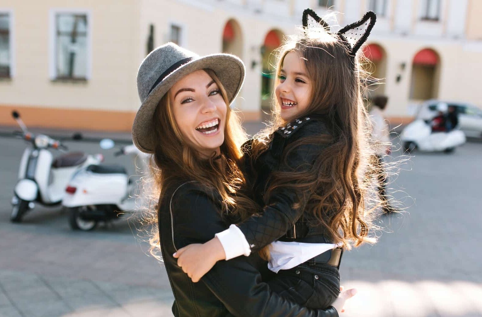Stylish young mom in retro felt hat carrying her cheerful curly brunette daughter across the street.